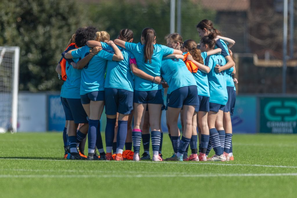 Worthing College team huddle at the Sussex County FA HQ before a friendly vs Chichester. They are wearing light blue shirts, navy blue shorts and socks and some people have bright orange bibs in their hand.