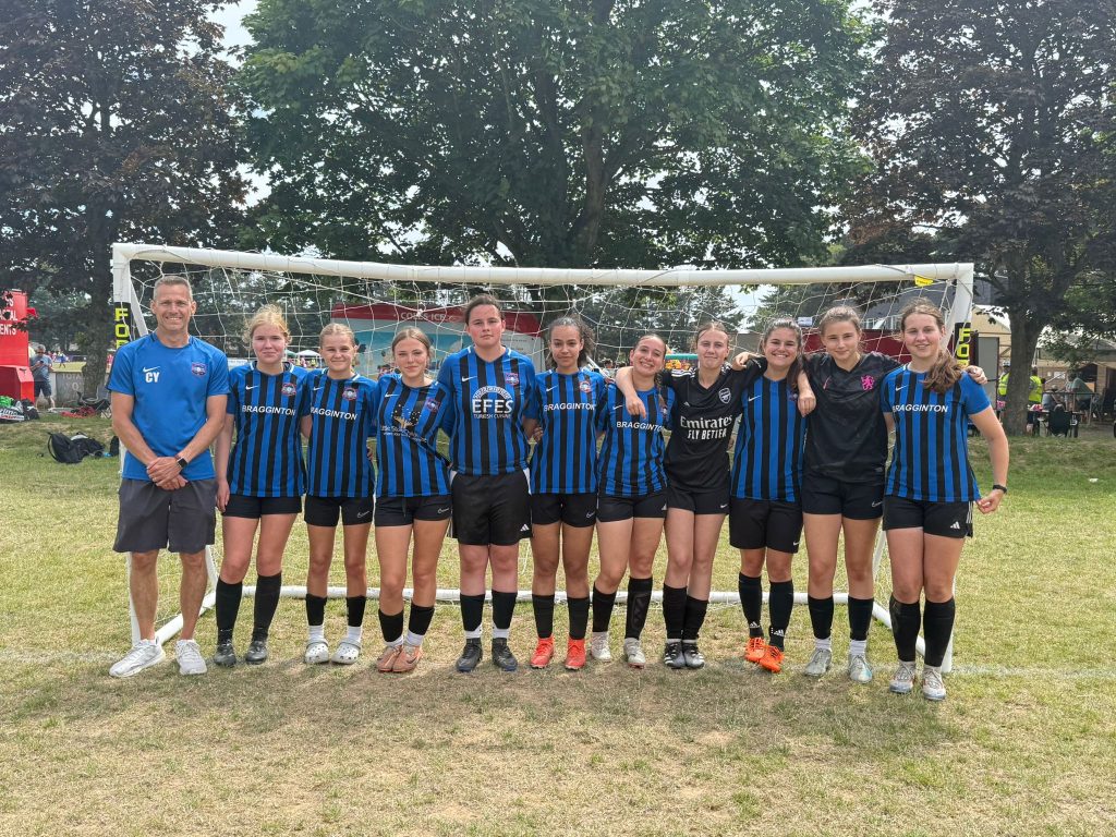 10 female football players and their coach in front of a 7-a-side goal at a tournament. They are wearing blue and black striped shirts and black shorts, some with sponsors some without. The goalkeepers are wearing full black kit