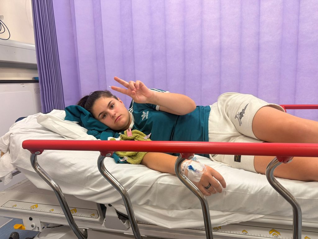 sad girl in teal adidas top and white shorts in hospital bed with a cannula doing a peace sign with her fingers. she is laying on her side looking at the camera